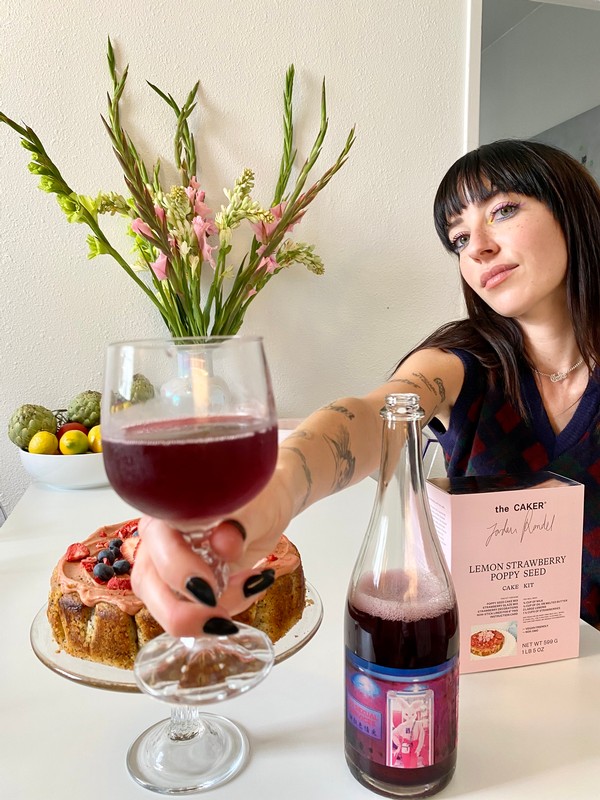 Jordan Rondel, The Caker, holding a glass of Tokyo Love Hotel to the camera. A Lemon Strawberry Poppyseed cake with mixed berries can be seen on the table.