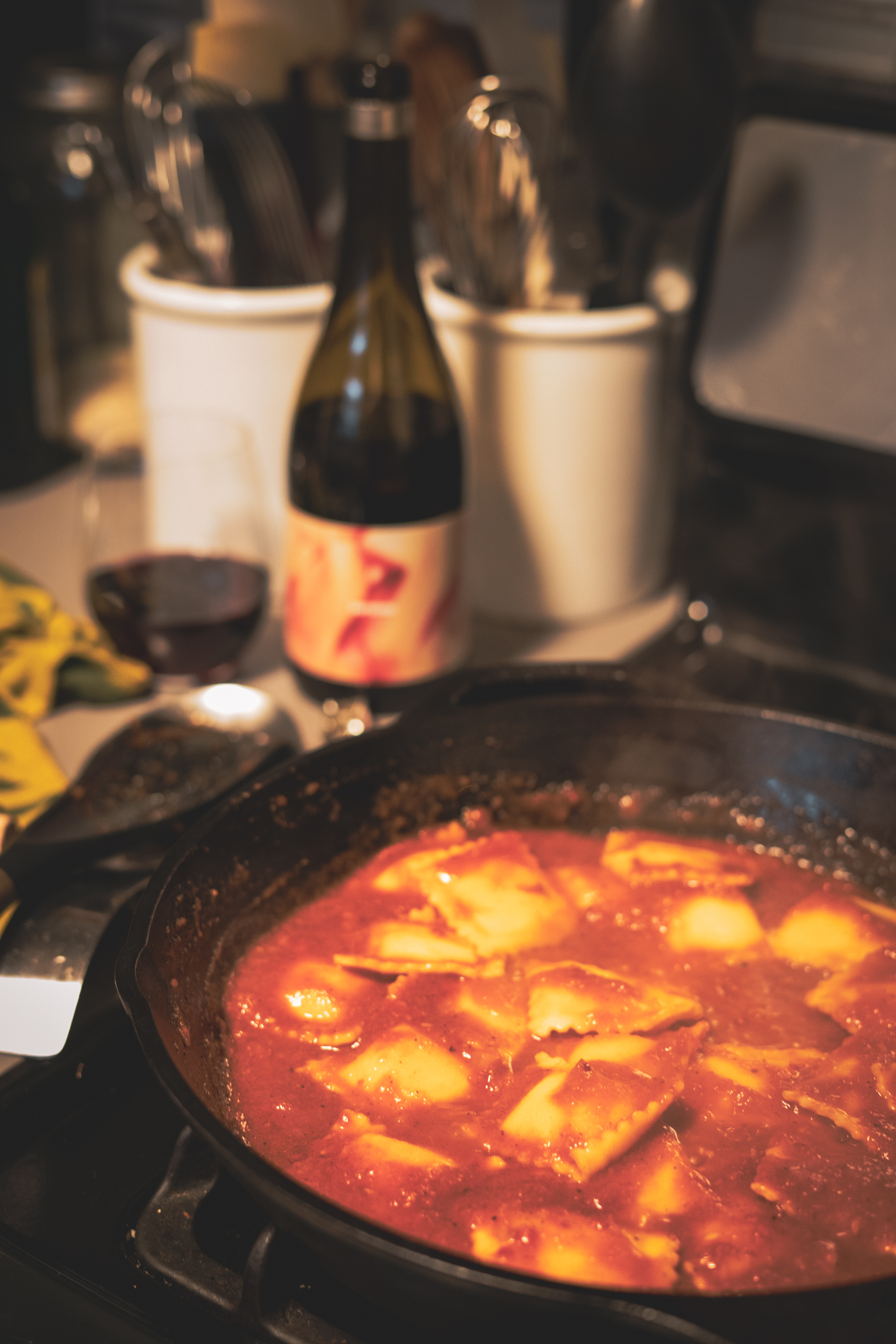 Ravioli with arrbiata sauce cooking on a stovetop in a cast iron pan. A bottle of Softcore wine is sitting on the counter top with a glass of wine next to it. There are two ceramic utensil bins behind the wine and the corner of a kitchen towel can be seen on the counter.