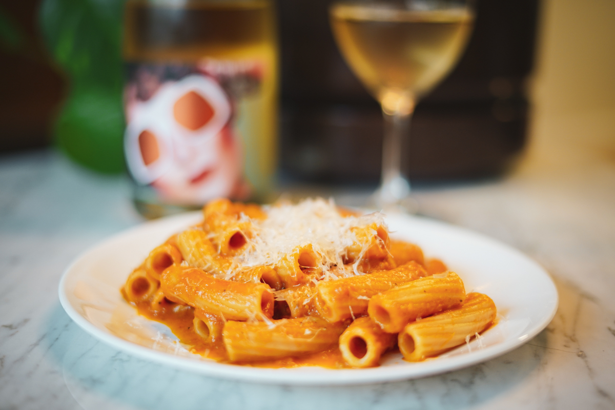A plate of pasta with roasted tomato sauce and a bottle and glass of Occhiali da Sole in the background.