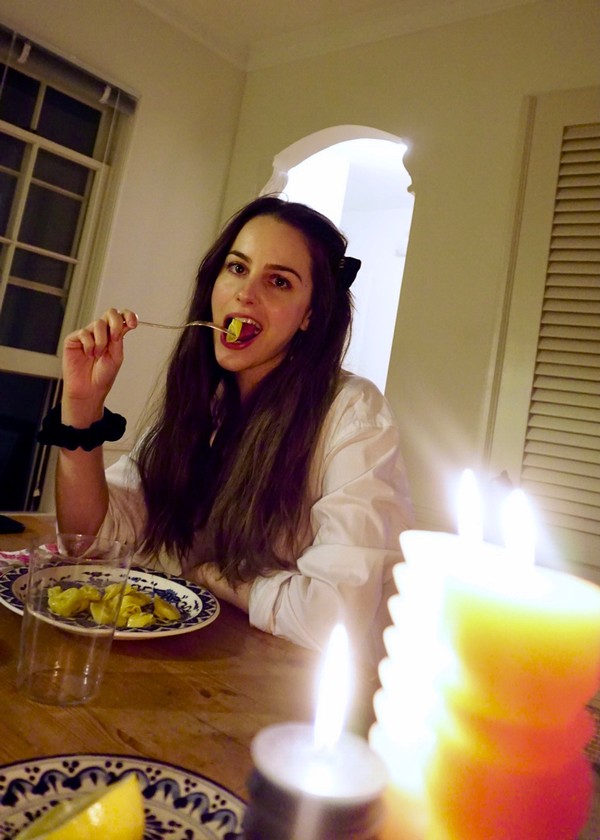 Rosie sitting at the dinner table in her home taking a bite out of a tortellini pasta. She has her hair down and is wearing a cuffed white shirt and a black scrunchie on her wrist.