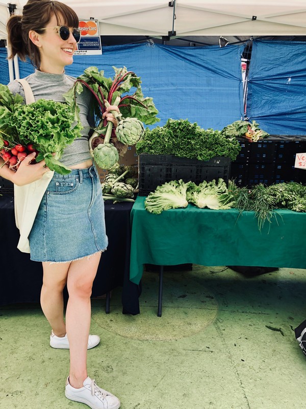 Rosie shopping at a local farmer's market, carrying beets and various greens. She's wearing a grey shirt with her hair pulled into a ponytail and wearing 60s inspired sunglasses.